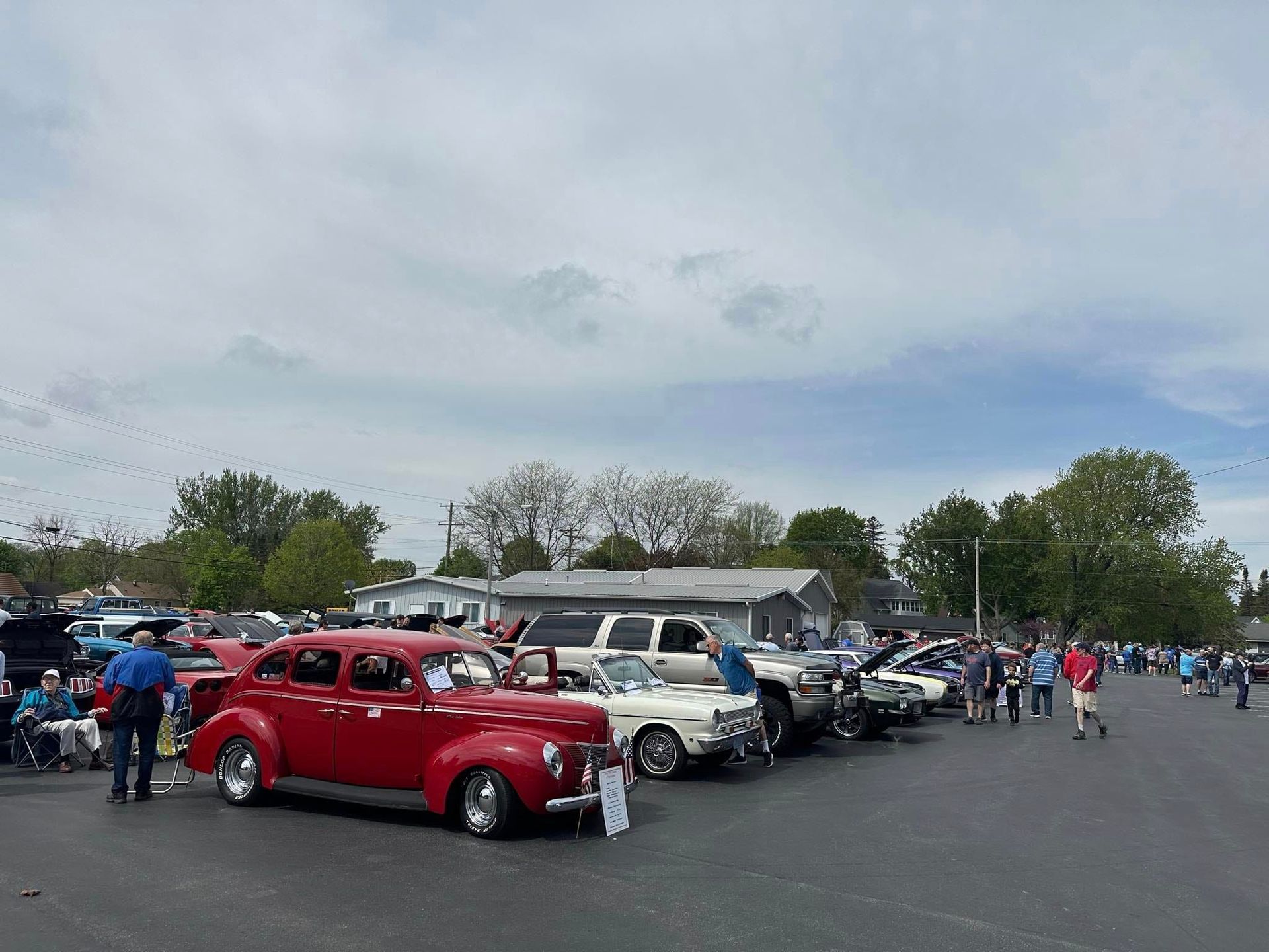 Classic cars parked at an outdoor car show on a cloudy day, with people walking around.