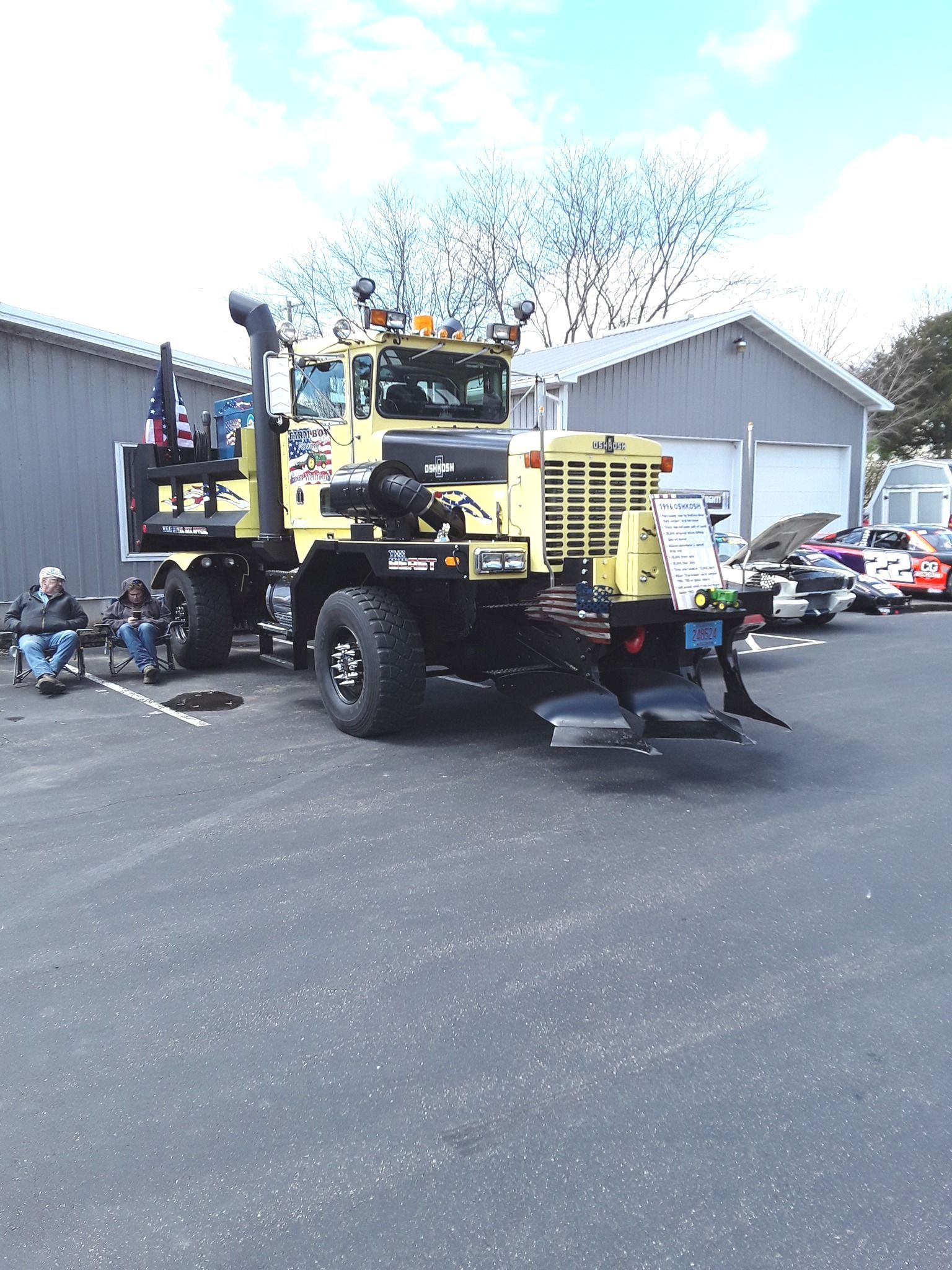 Yellow heavy-duty truck parked on asphalt. People sitting nearby. Buildings in the background under a blue sky.