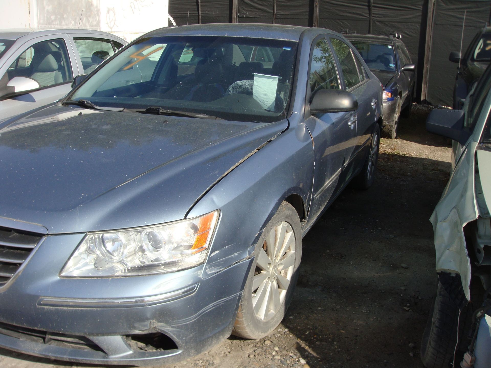 Blue sedan parked in a lot beside other cars, with a damaged front bumper and headlight