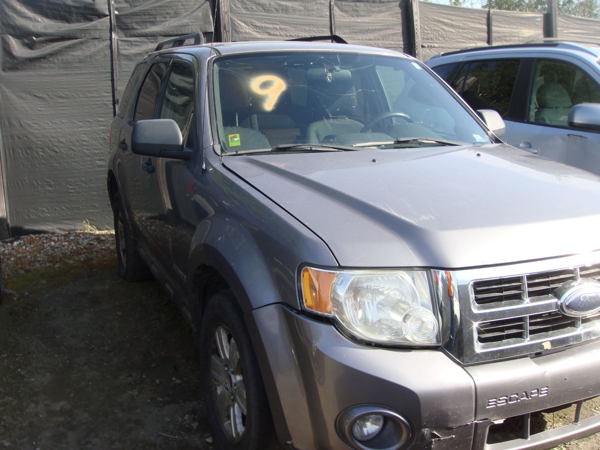 Gray SUV parked outdoors beside a tarp wall, with a yellow “9” on the windshield.