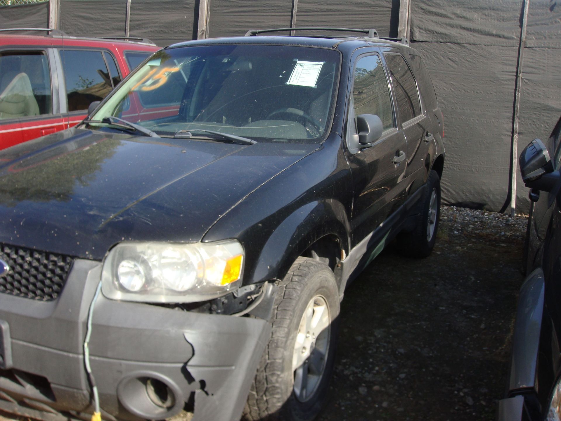Black SUV parked in a lot, front bumper damaged and headlight fogged, next to a red car and wall.