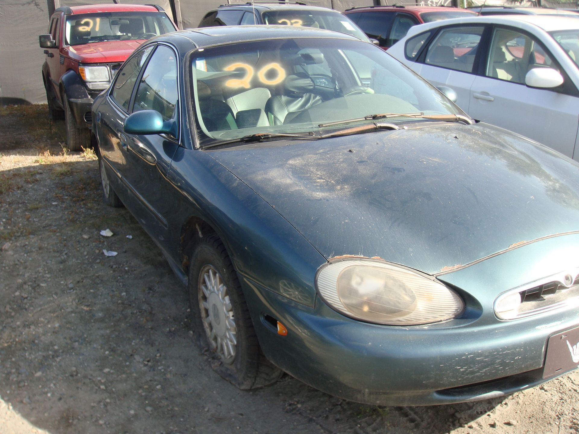 Blue sedan with yellow “20” on the windshield, parked in a crowded lot behind a red car.