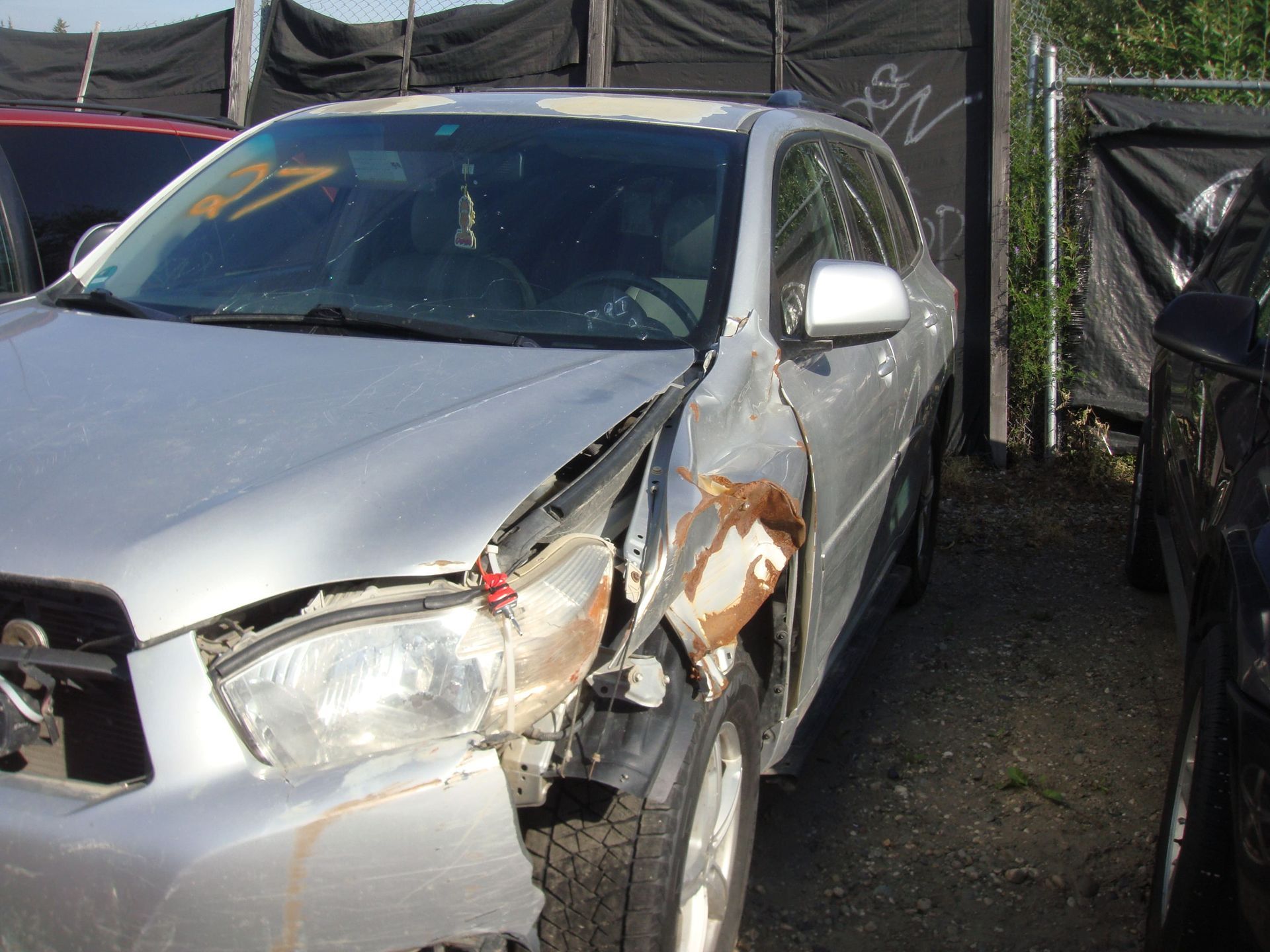 Silver car with severe front-left body damage in a lot, next to a black tarp fence.
