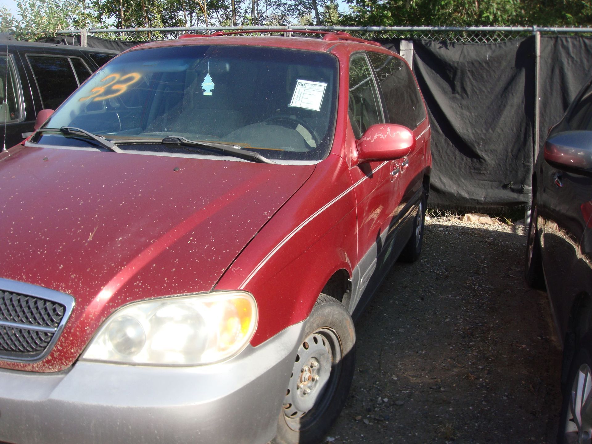 Red minivan parked outdoors next to a covered vehicle on gravel.