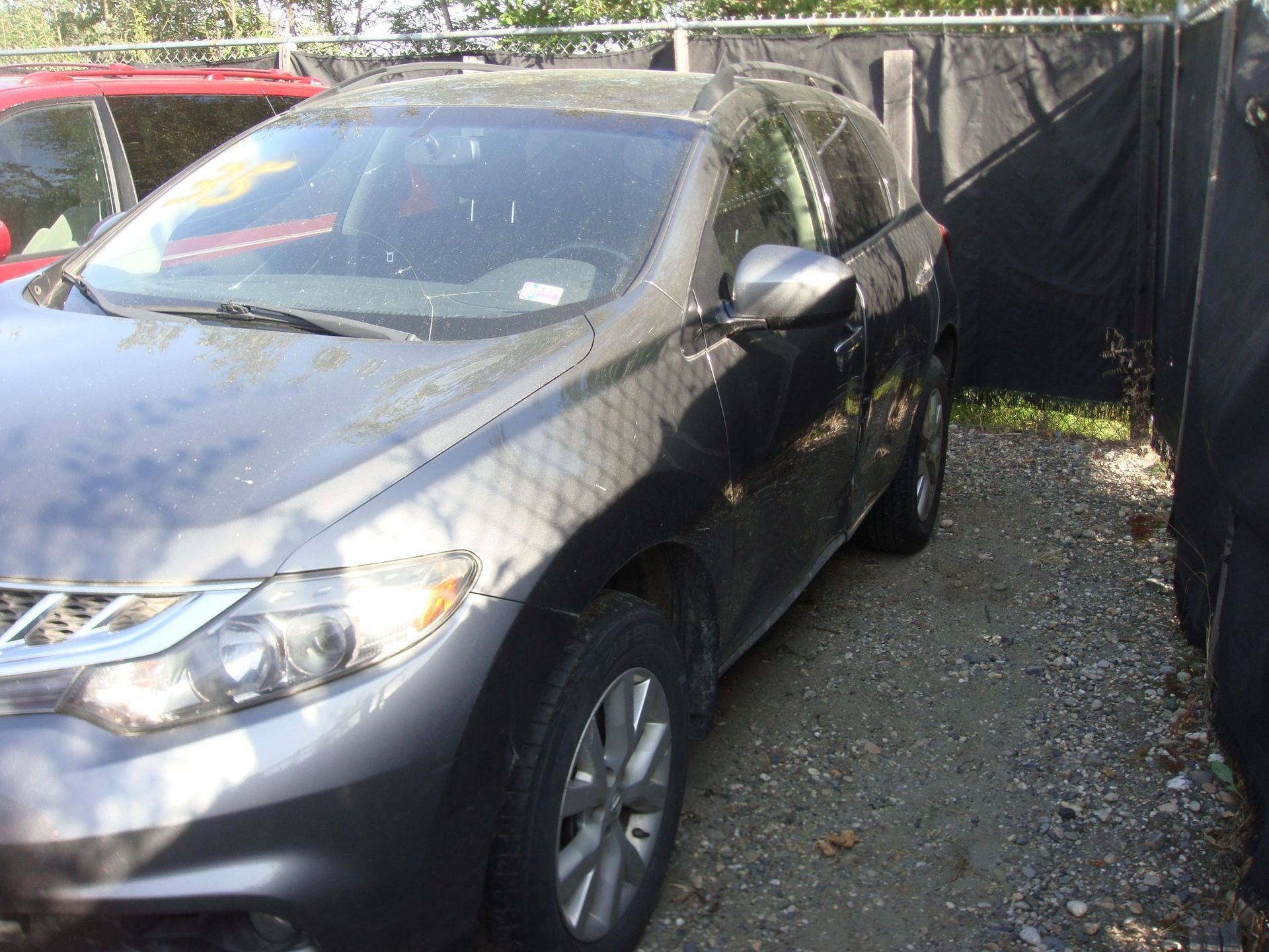 Gray SUV parked in a narrow gravel driveway beside a black fence and red car