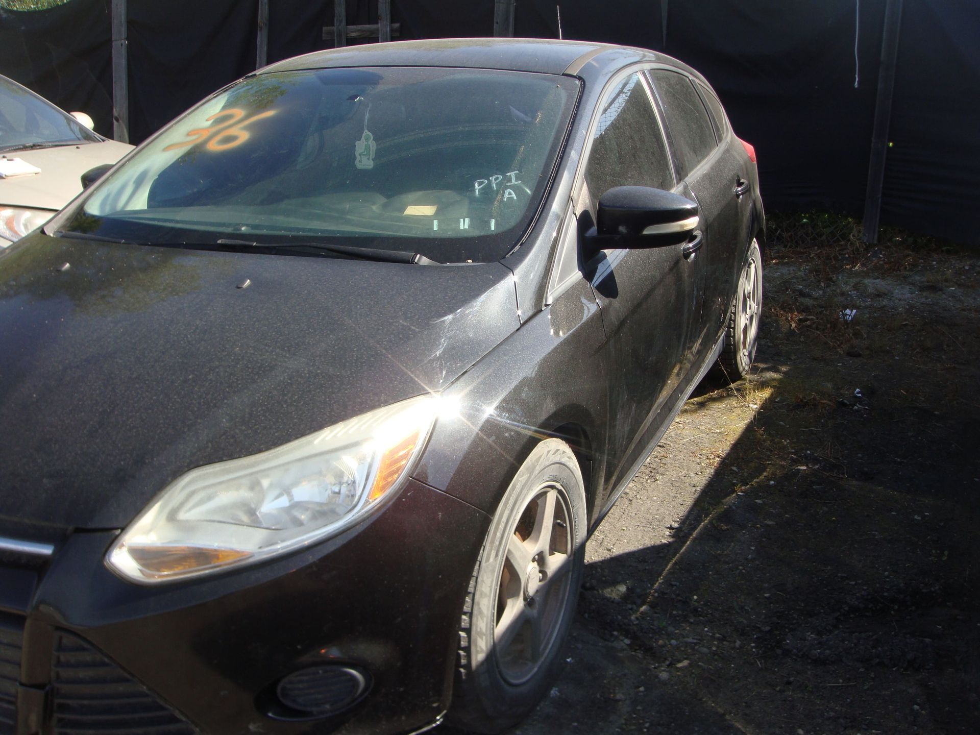 Dark gray sedan parked outdoors beside a black backdrop, with sunlight on the hood.