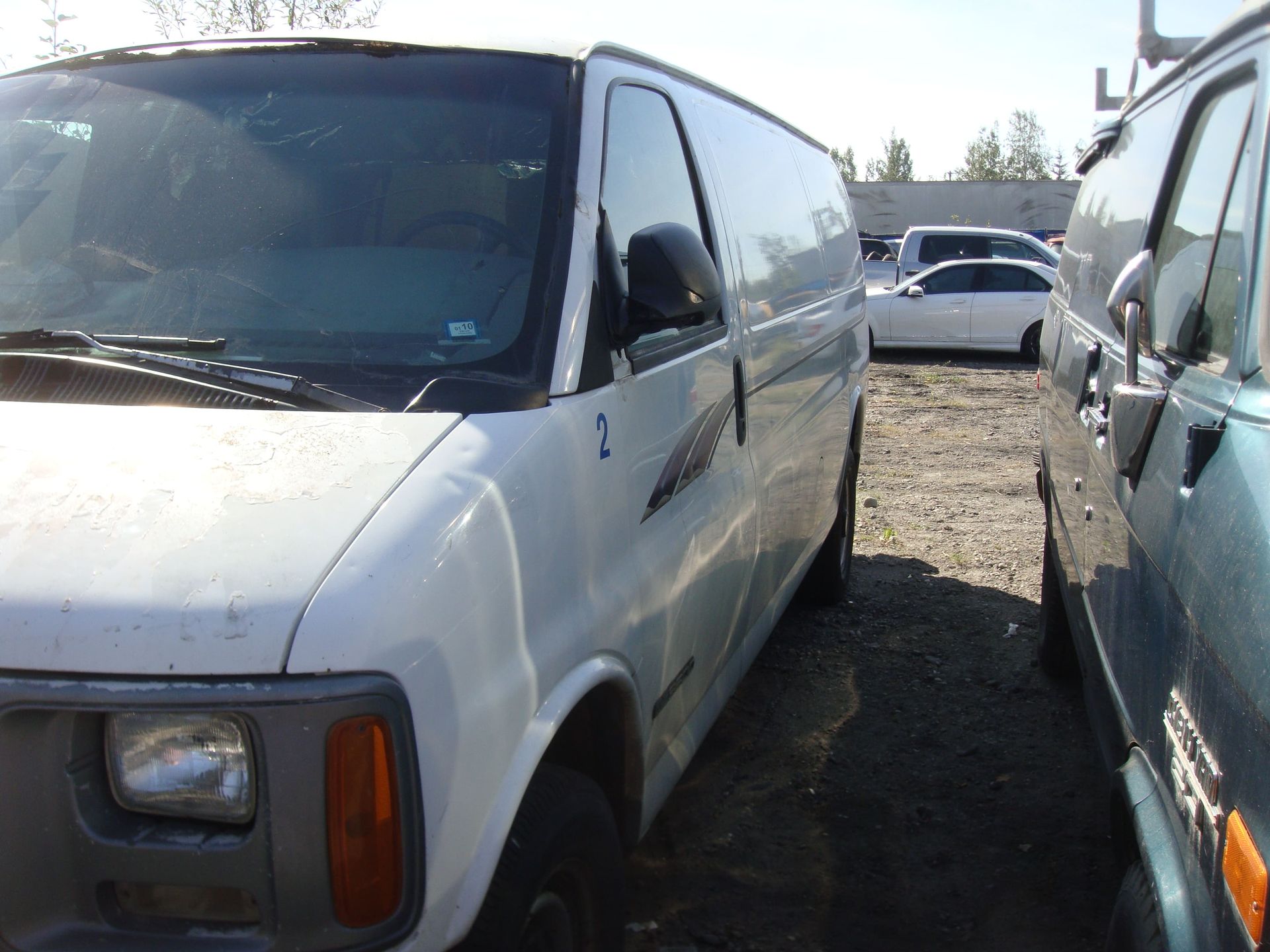 White cargo van parked in a gravel lot beside a blue van, with another vehicle in the background