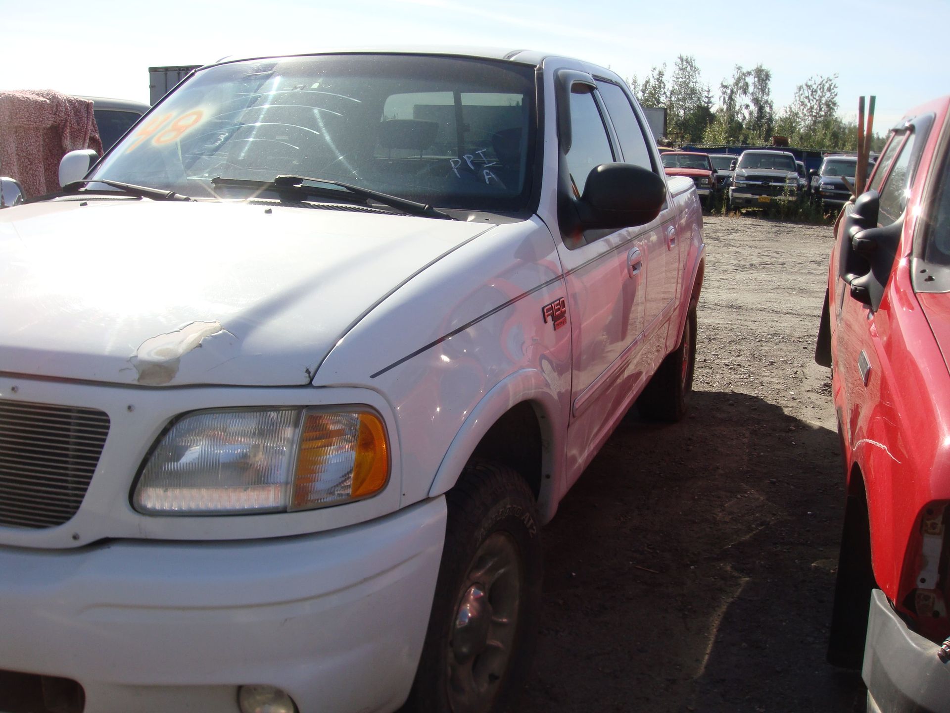 White pickup truck in a salvage yard, parked beside a red vehicle under bright sunlight