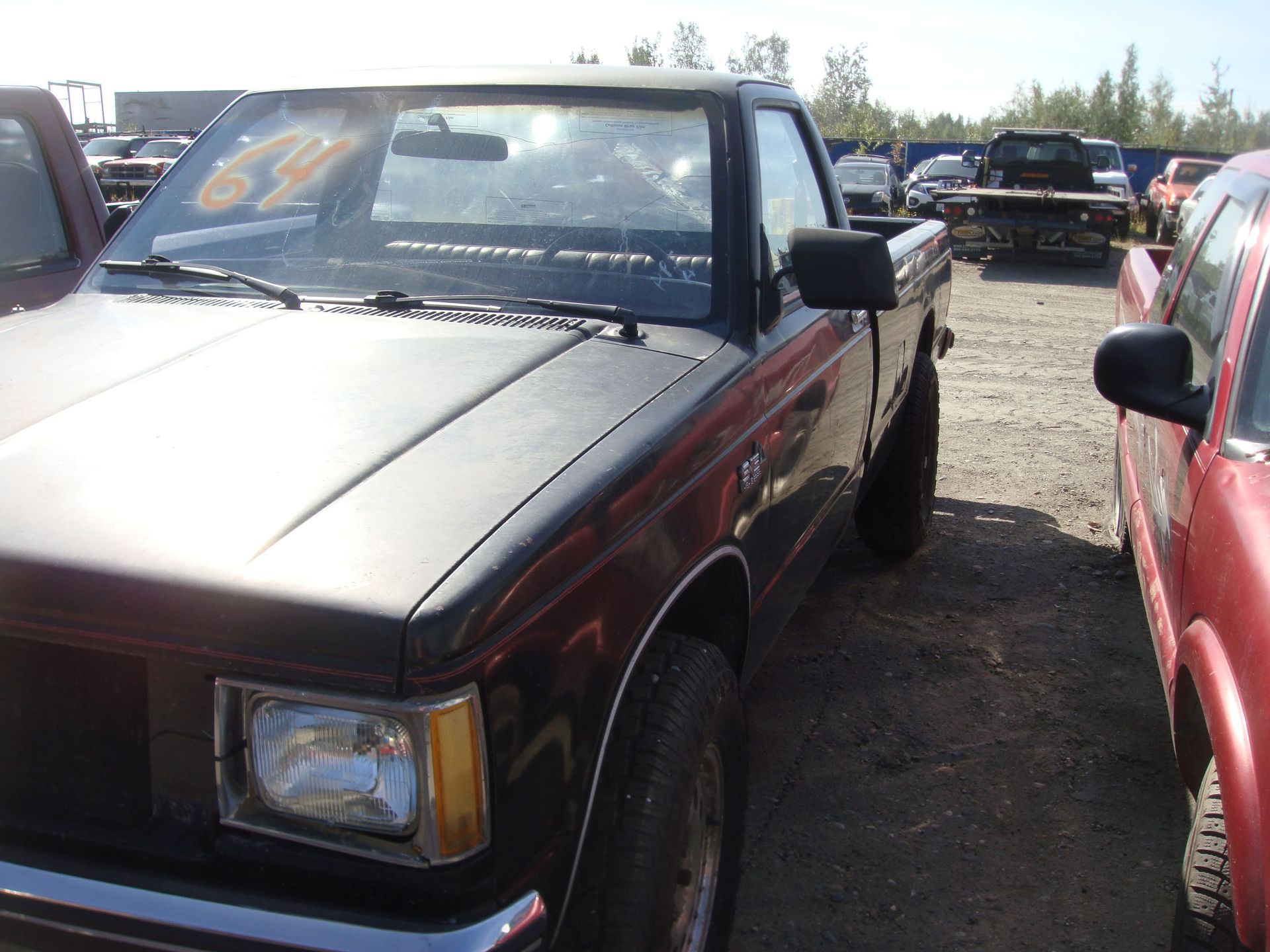 Red pickup truck parked in a dirt lot beside other vehicles under bright sunlight.