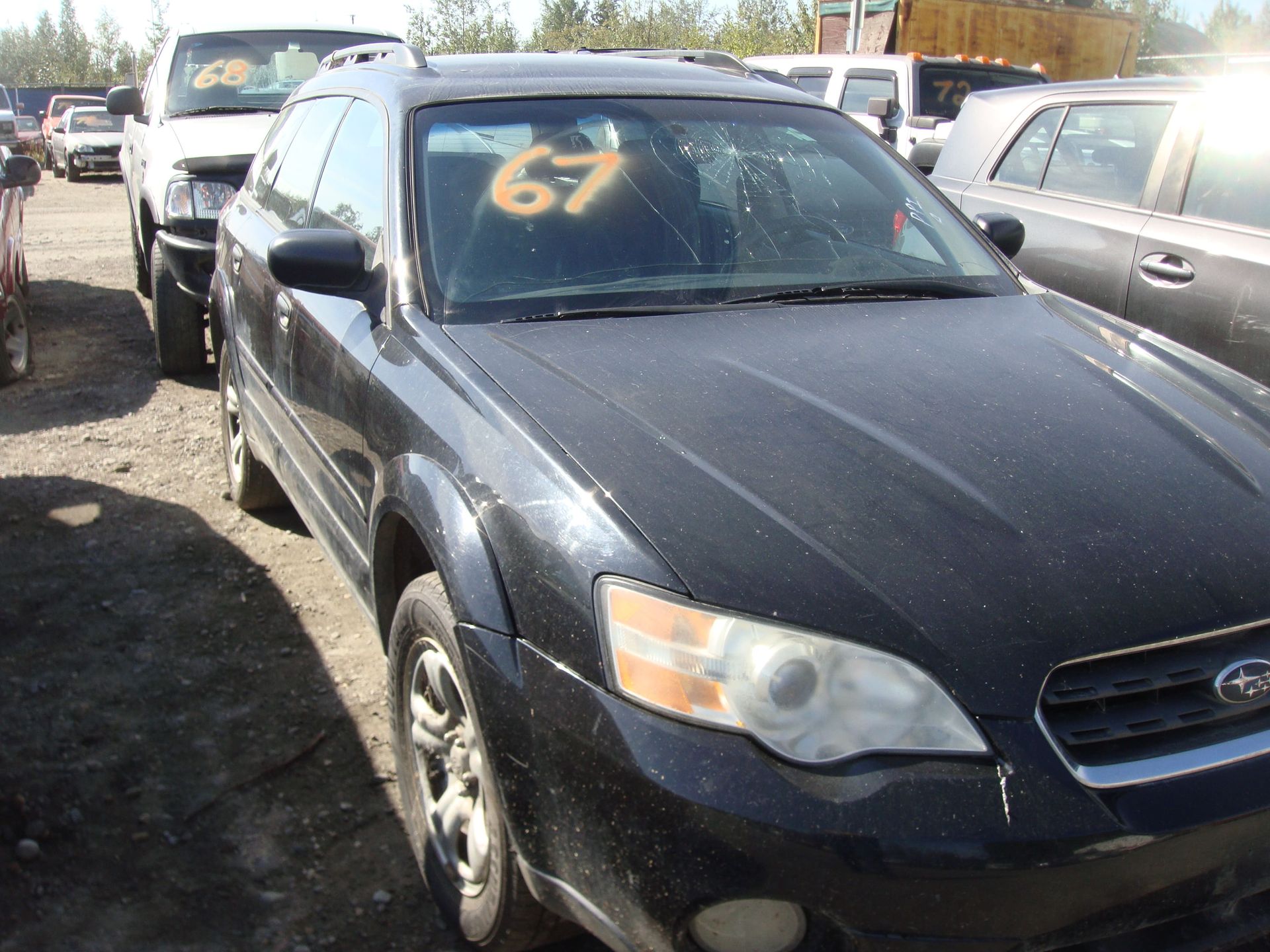 Black sedan in a car lot with orange “67” on the windshield, parked beside other vehicles.