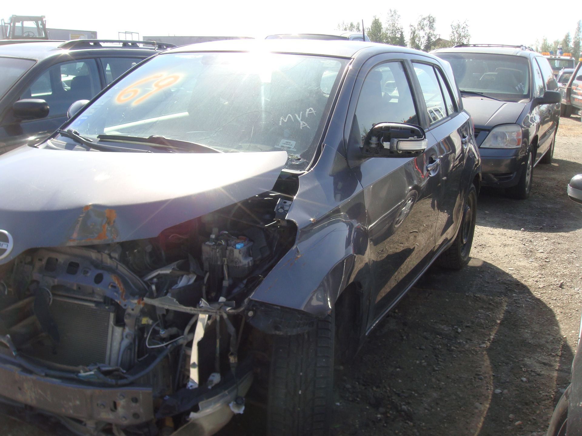Damaged black hatchback with crushed front end in a junkyard, parked among other wrecked cars