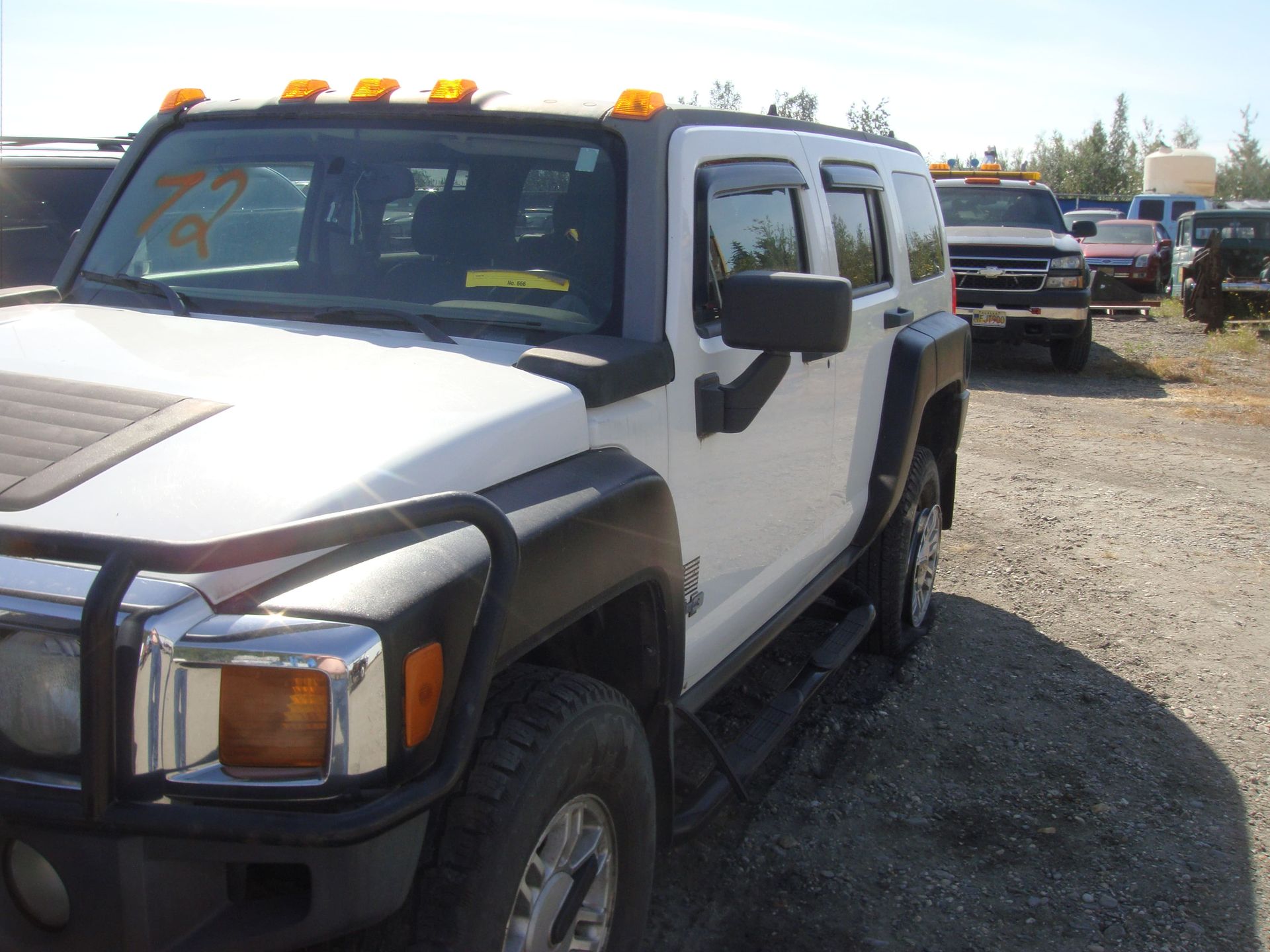 White Hummer H2 parked on a gravel lot with amber roof lights and a chrome grille guard.