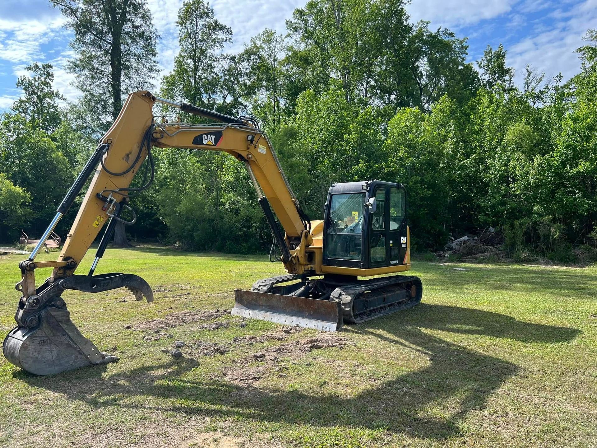 A yellow Caterpillar hydraulic excavator sits on a grassy field in front of a dense treeline on a sunny day.