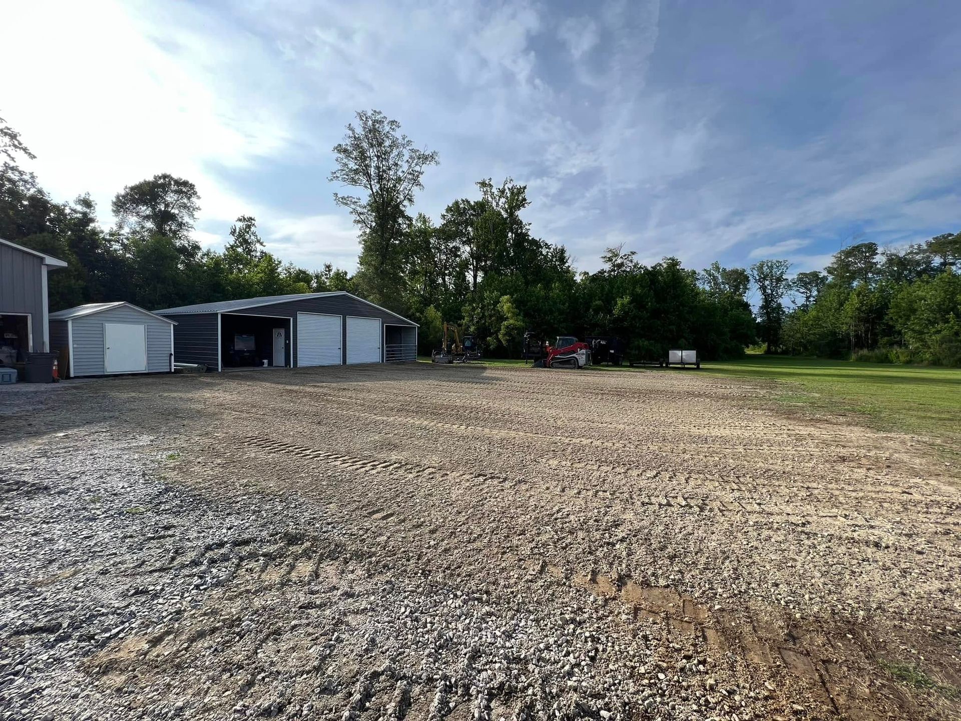 A gravel lot leads to two metal outbuildings in front of a treeline under a blue, cloudy sky.