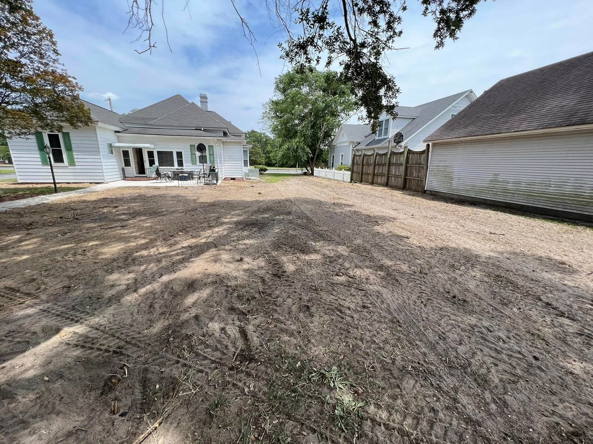 A dirt backyard under a bright sky, featuring a white house on the left and a neighboring building with a fence.