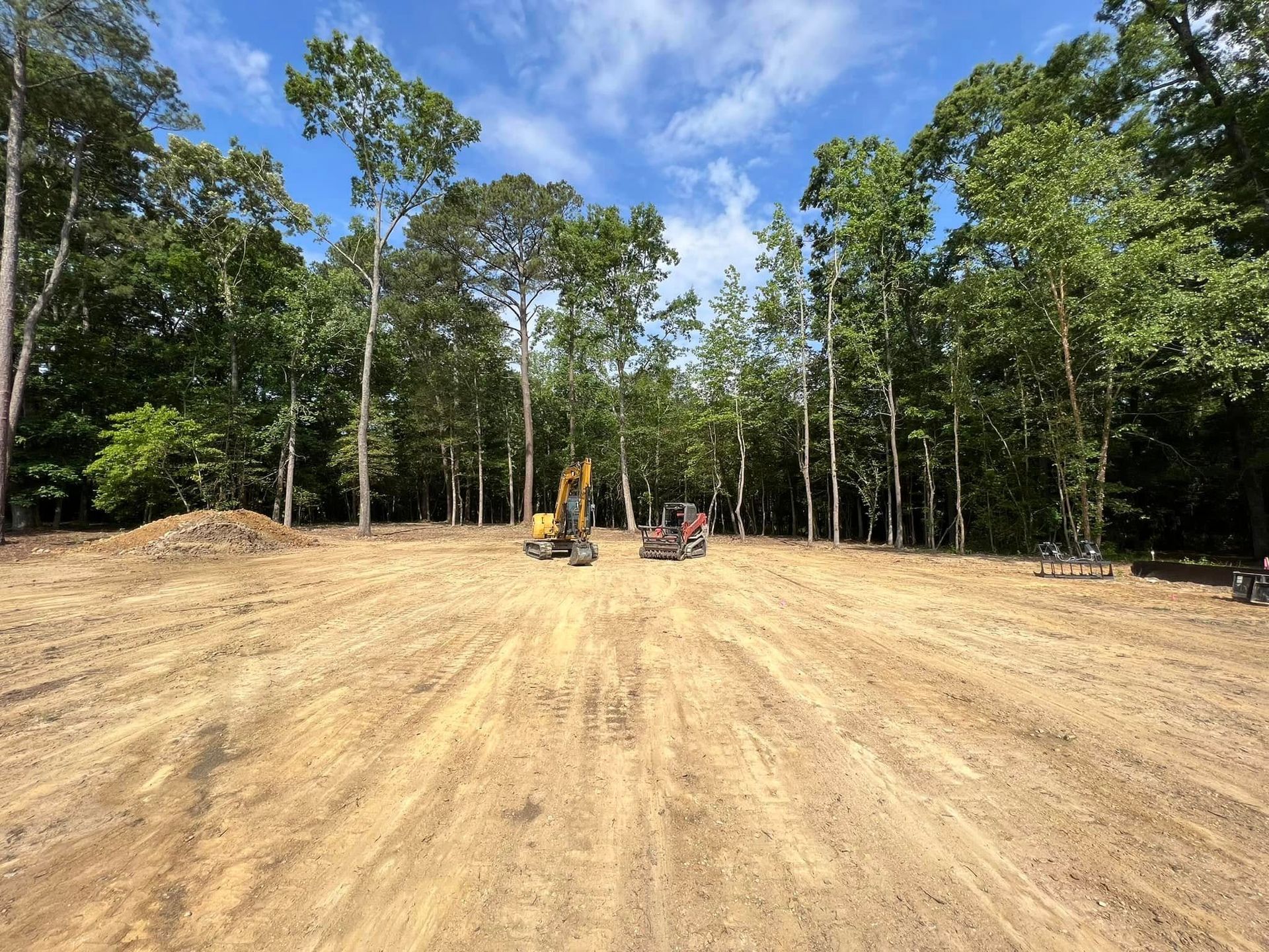 Two pieces of yellow construction equipment sit on a cleared dirt lot bordered by a dense forest under a blue sky.