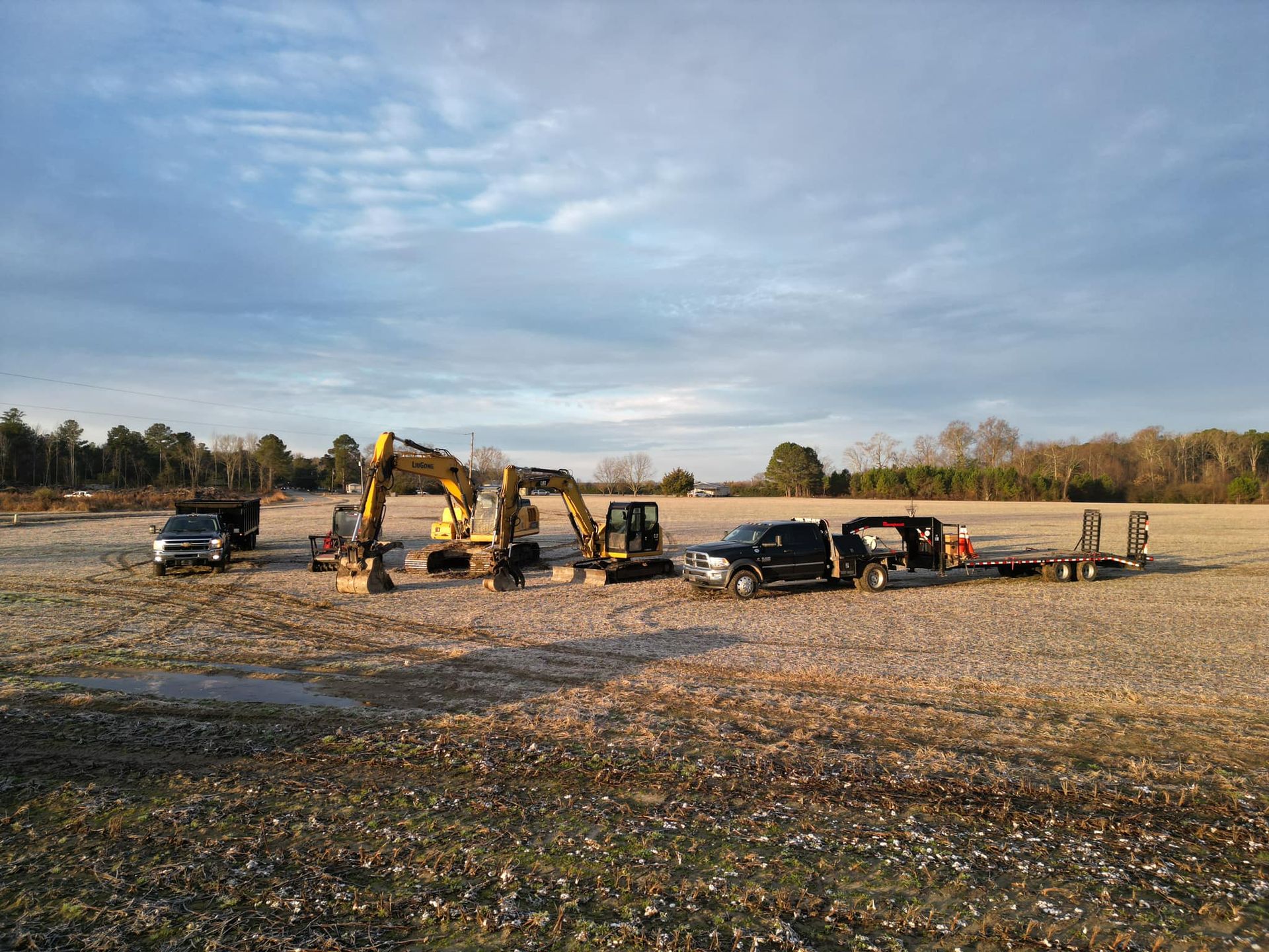 Excavators and pickup trucks lined up on a rocky riverbed under a cloudy sky, with workers nearby.