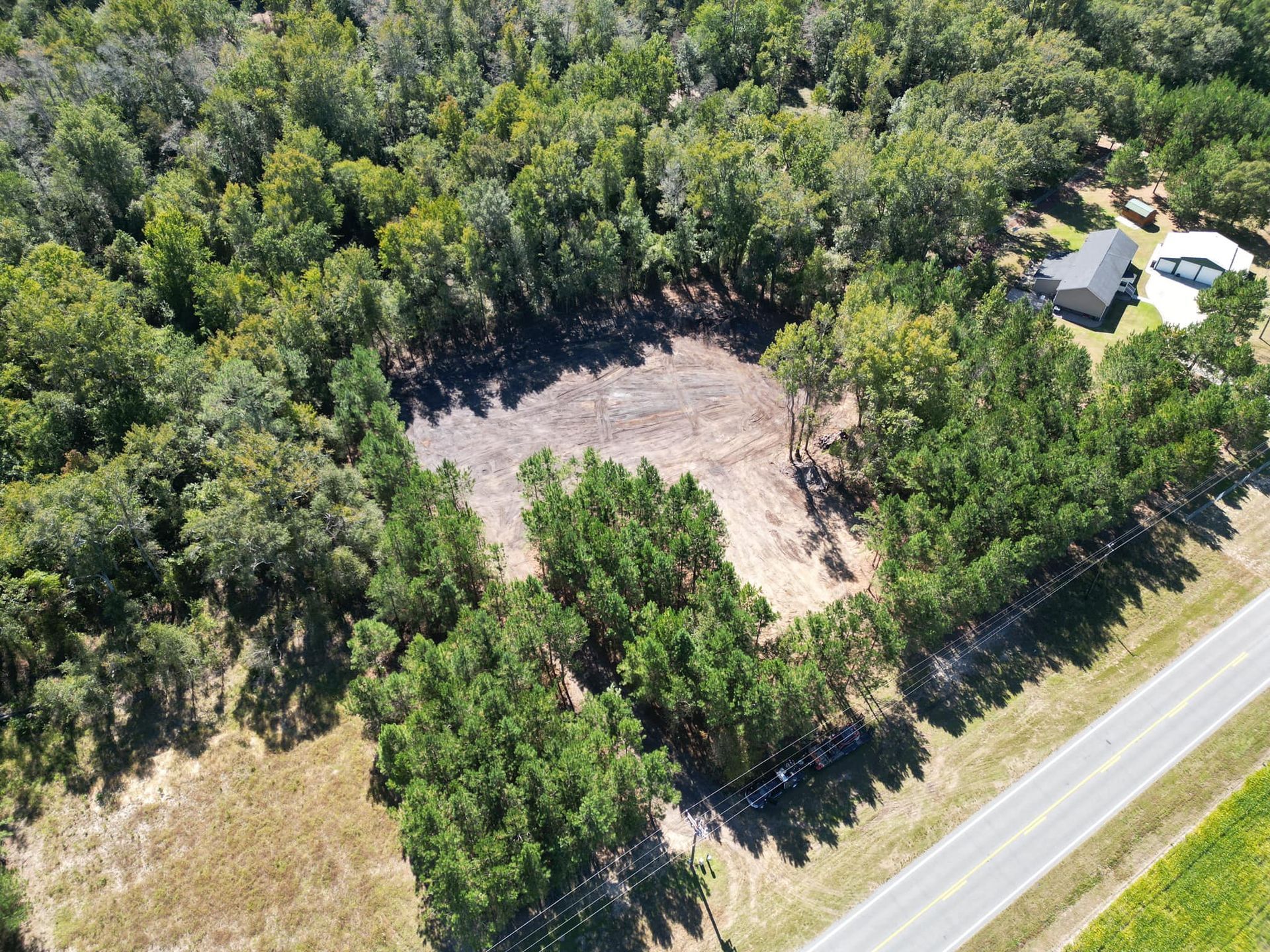 An aerial view of a cleared patch of land surrounded by trees next to a road and a nearby house.