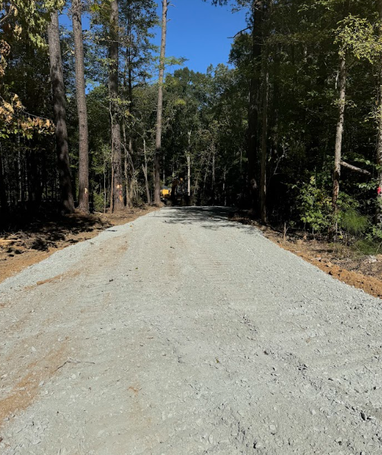 Gravel road under construction through a forest, with tall trees on both sides and a clear blue sky.