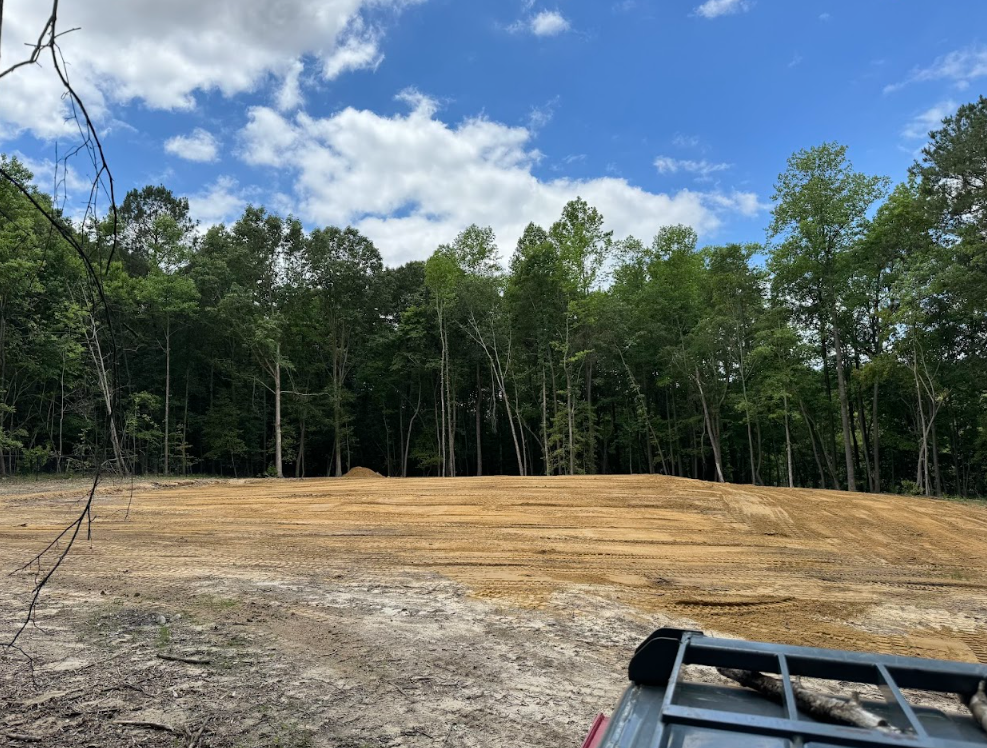 Cleared dirt lot beside a wooded tree line under a blue sky, with part of a vehicle visible in the foreground