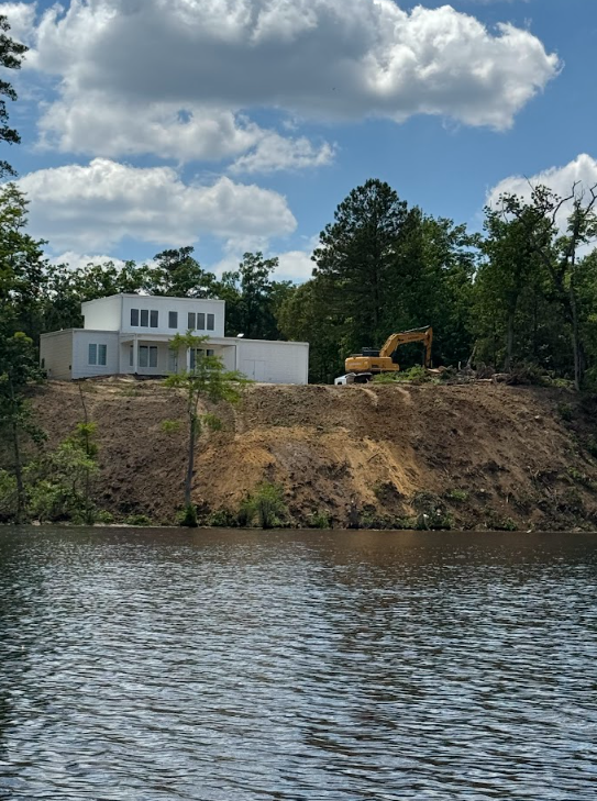 Riverside home under construction with an excavator on a dirt bank under a cloudy sky