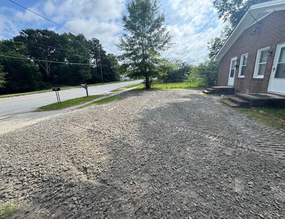Gravel yard beside a brick building, with a road, trees, and cloudy sky in the background
