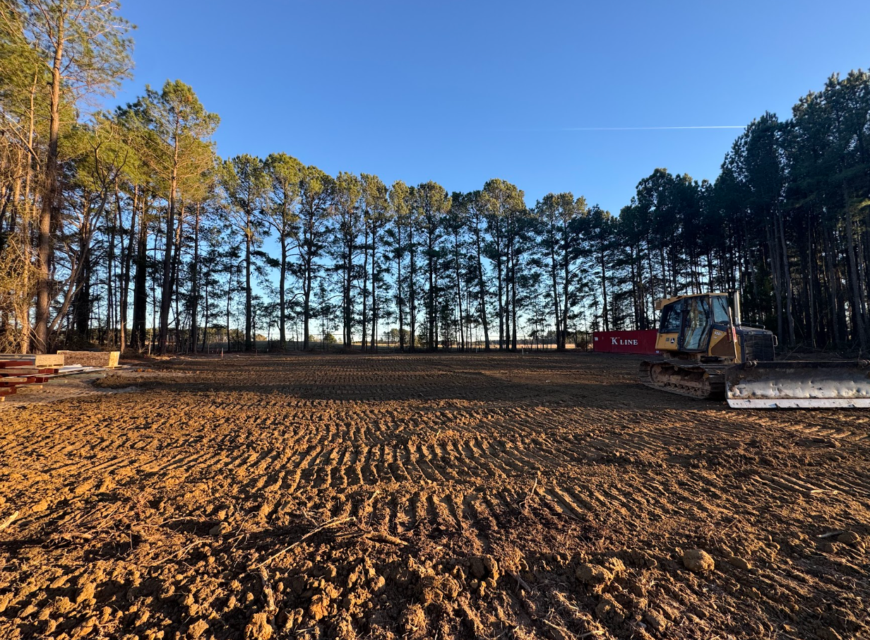 Cleared dirt lot bordered by tall trees under a blue sky at sunset