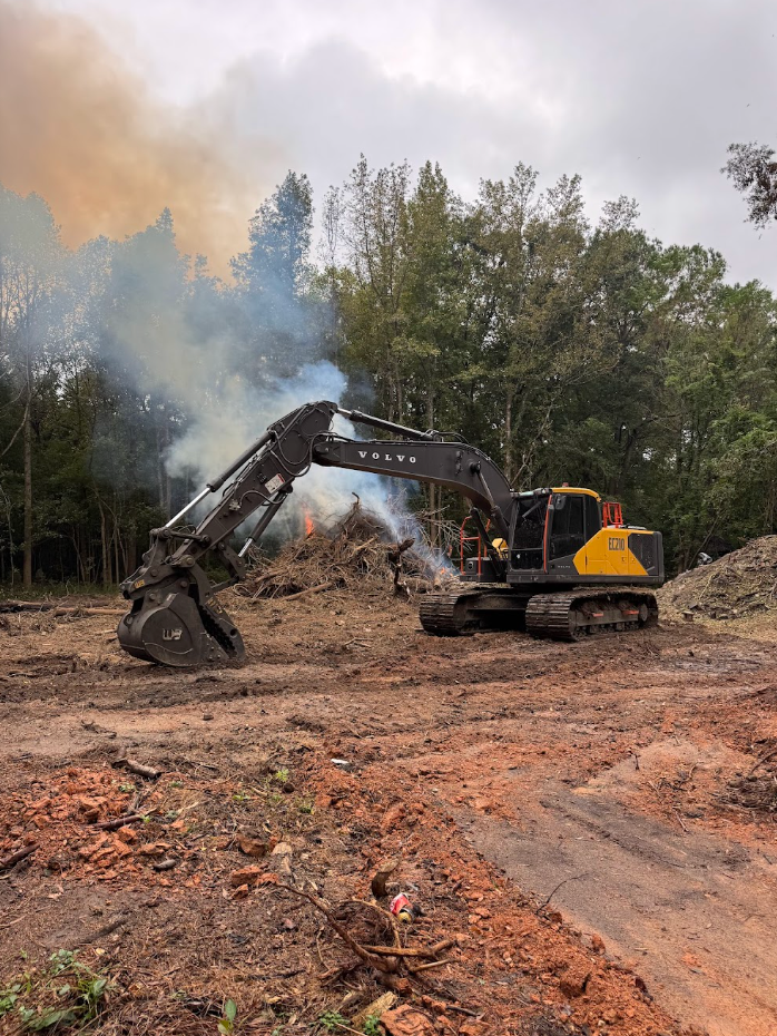 Excavator clearing debris and brush amid smoke in a muddy wooded area