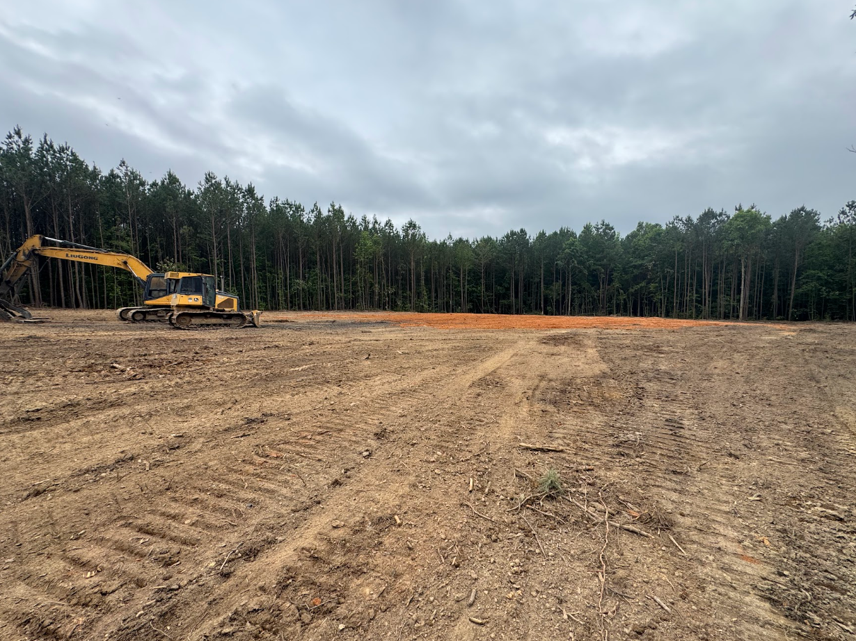 Excavator on a cleared dirt site bordered by trees under a cloudy sky