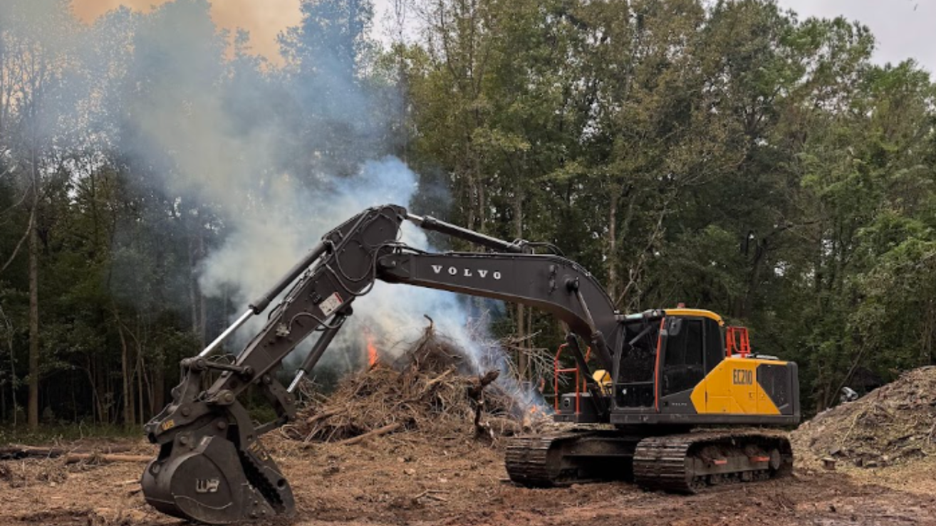 Excavator demolishing a brush pile with smoke rising in a wooded clearing