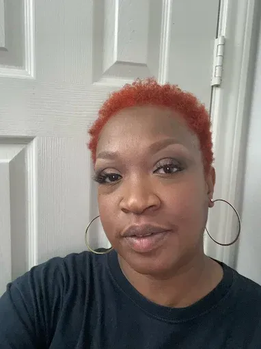 Close-up portrait of a person with short red hair and hoop earrings, standing by a white door.