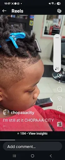 Child with braided hair and blue clips watching a phone screen with social media video controls visible.