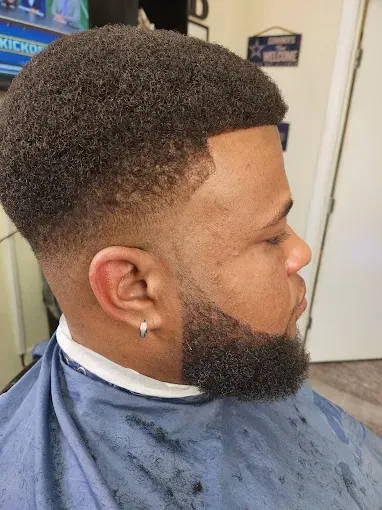 Side-profile of a man with a fresh fade haircut and beard, seated in a barber chair with a dark cape.