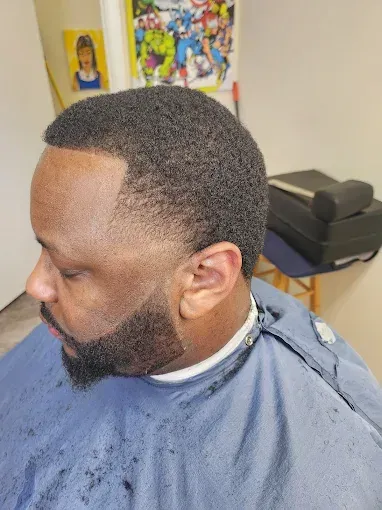 Side-profile of a man with a close-cropped fade and beard, seated in a barber chair wearing a blue cape.