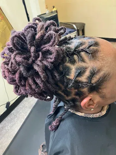 Person with purple locs in an intricate updo, showing a braided scalp design and gold jewelry.