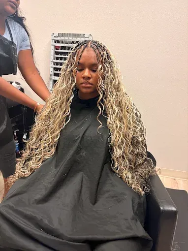 Person seated in a salon chair with long blonde braids, wearing a black cape, with another person in the background