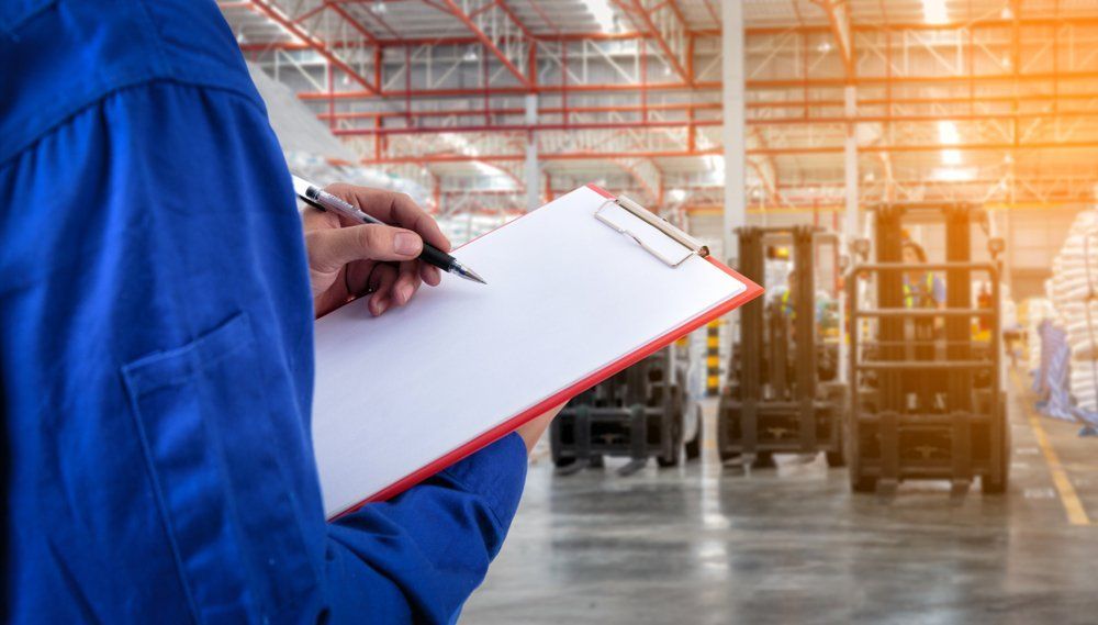 Person In Blue Jumpsuit With Clipboard And Pen In A Warehouse With Forklifts — Wendy's Safety Services in Moulden, NT