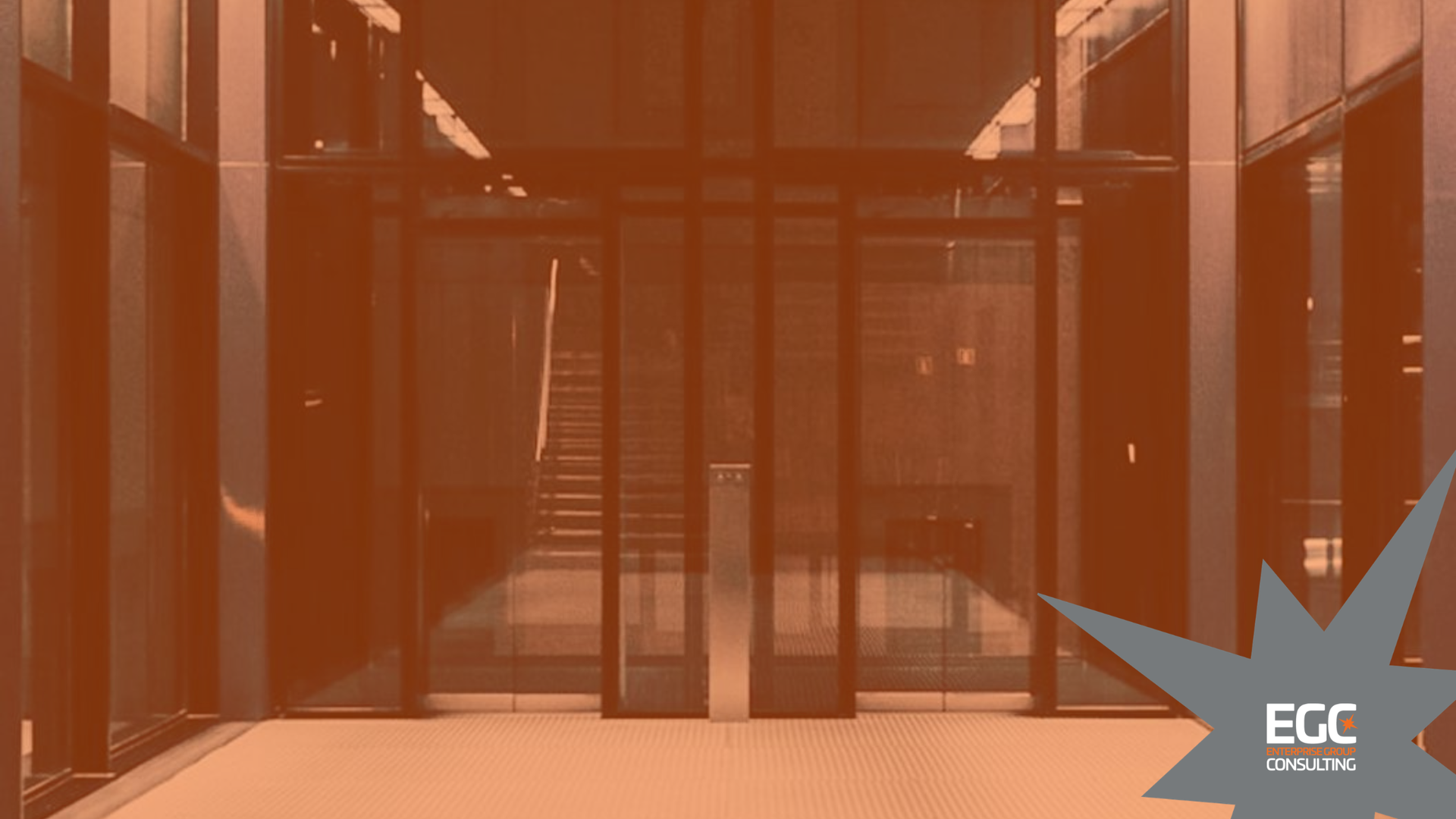 Glass-fronted building entrance with metal frame, seen through a warm orange tint, with a small logo in the corner