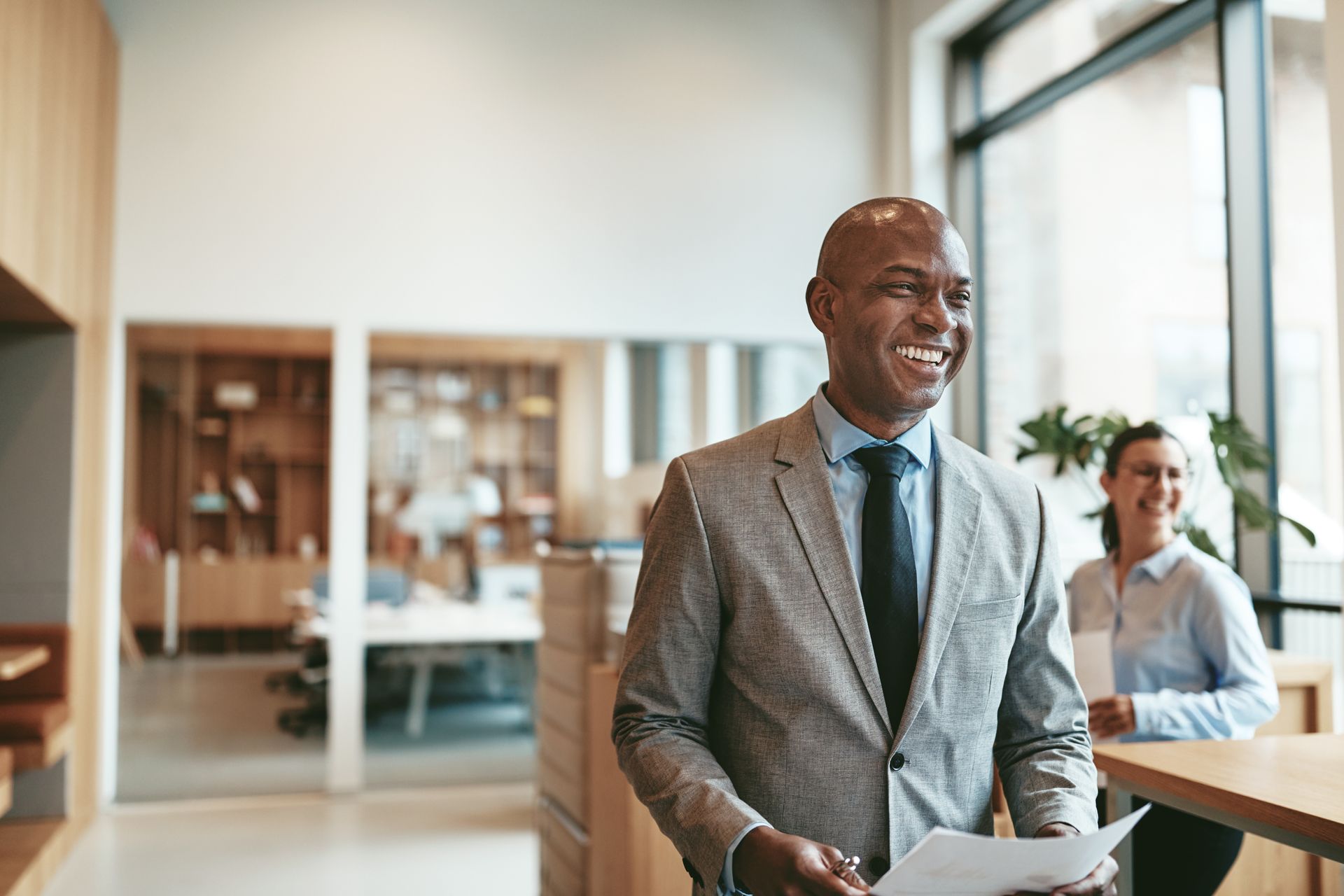 Man in suit smiles, holding papers, walking in office. Woman in background.