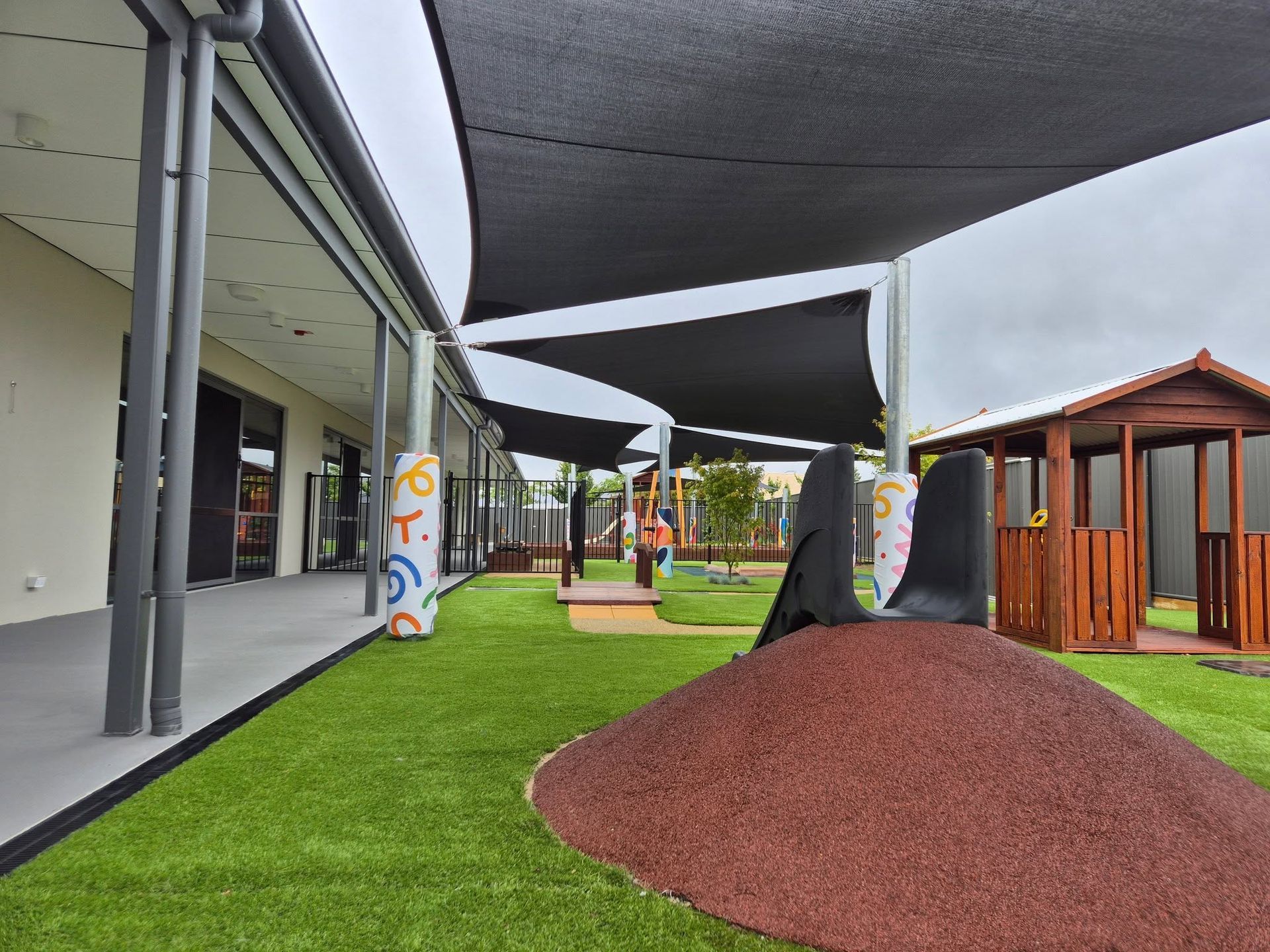 Modern playground with shade sails, green turf, red mulch mounds, and wooden playhouse structures
