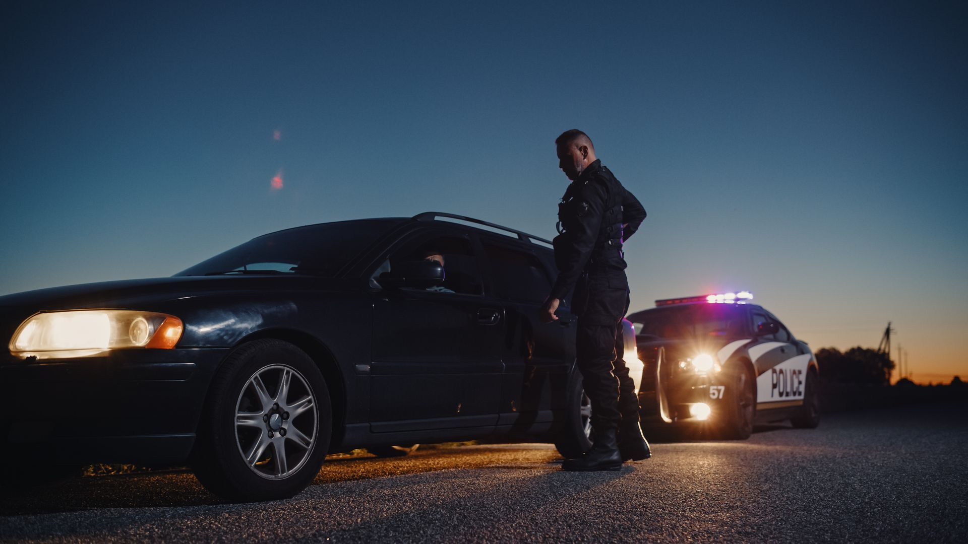 Police officer by a stopped car at dusk, patrol car lights flashing.