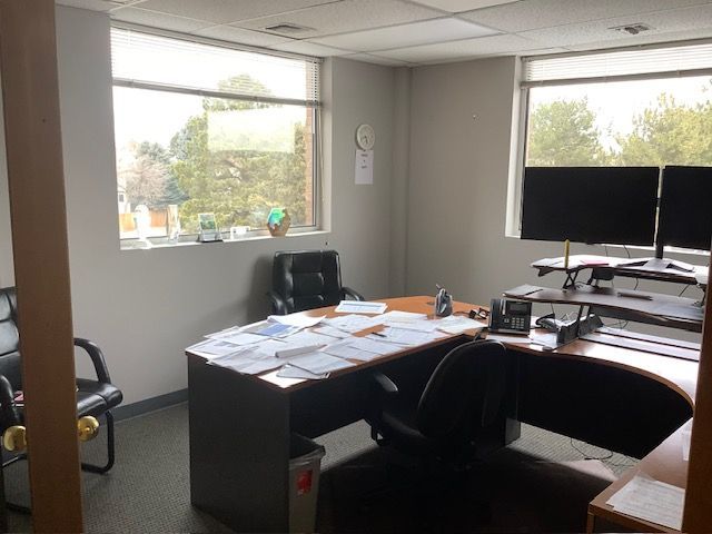 Empty office with desk clutter, dual monitors, and large windows overlooking trees.