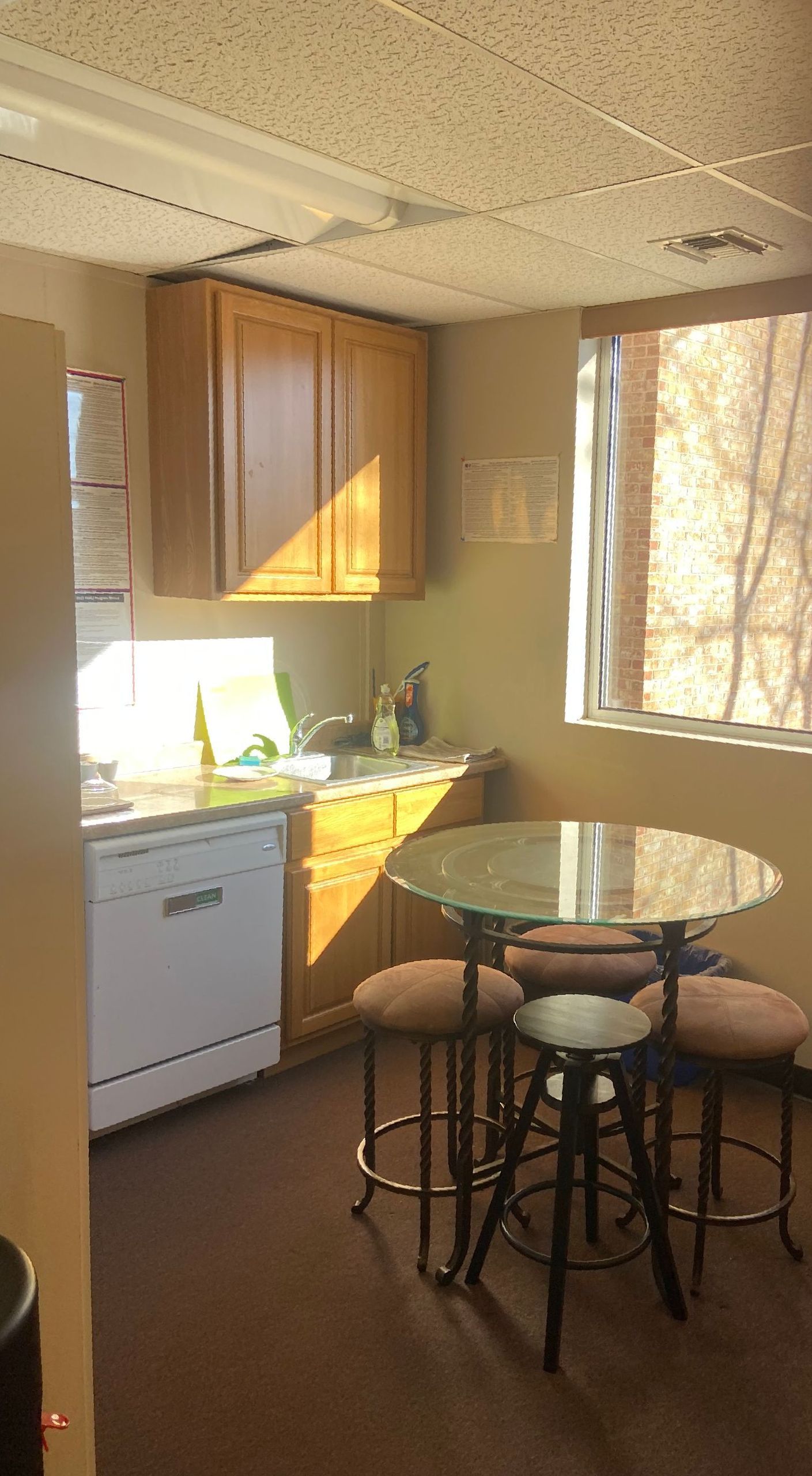 Small kitchen with wood cabinets, white appliances, a round glass table, and stools by a sunlit window.