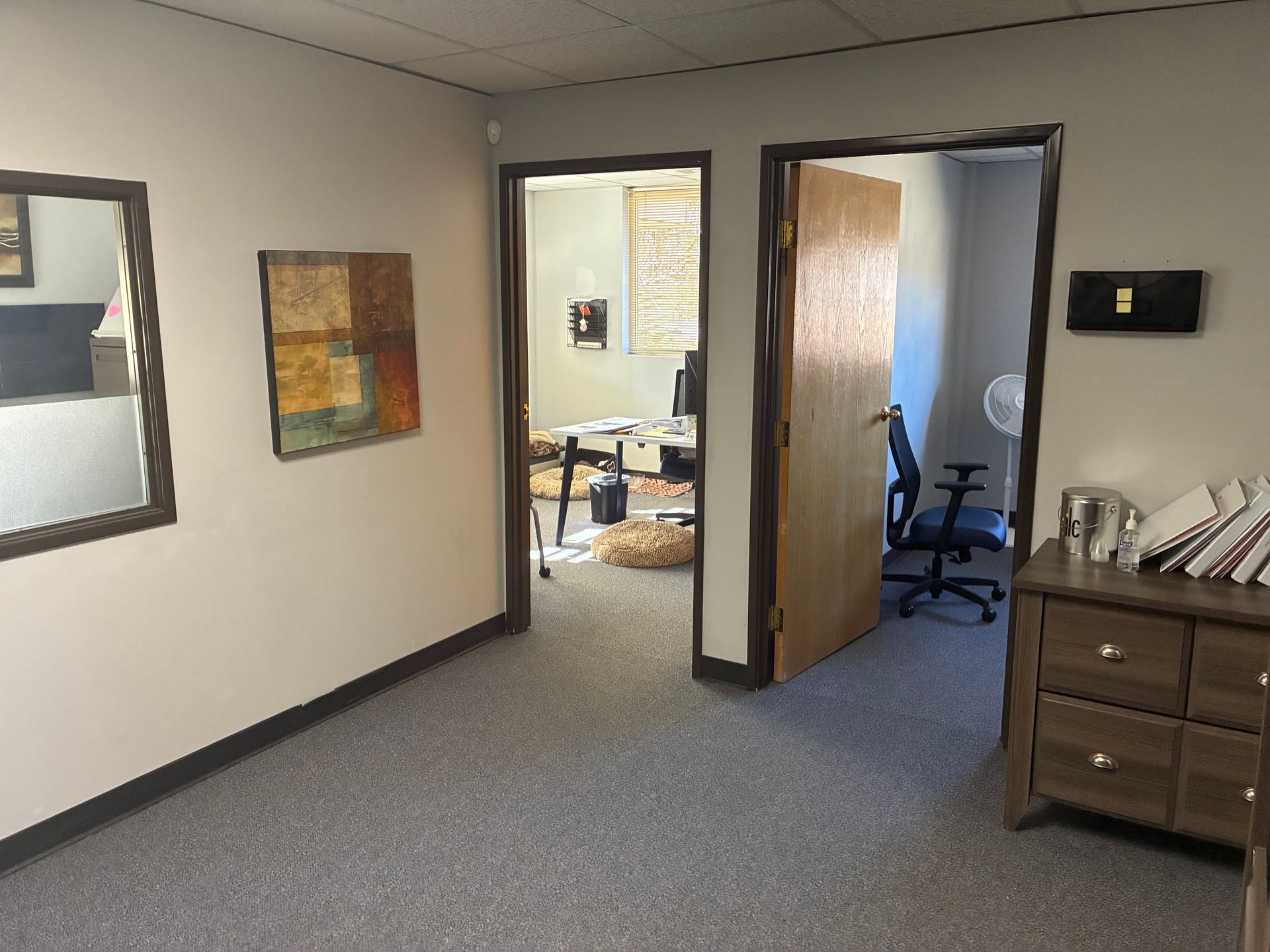Empty office hallway with carpet, open doors, wall art, and a wooden dresser on the right.