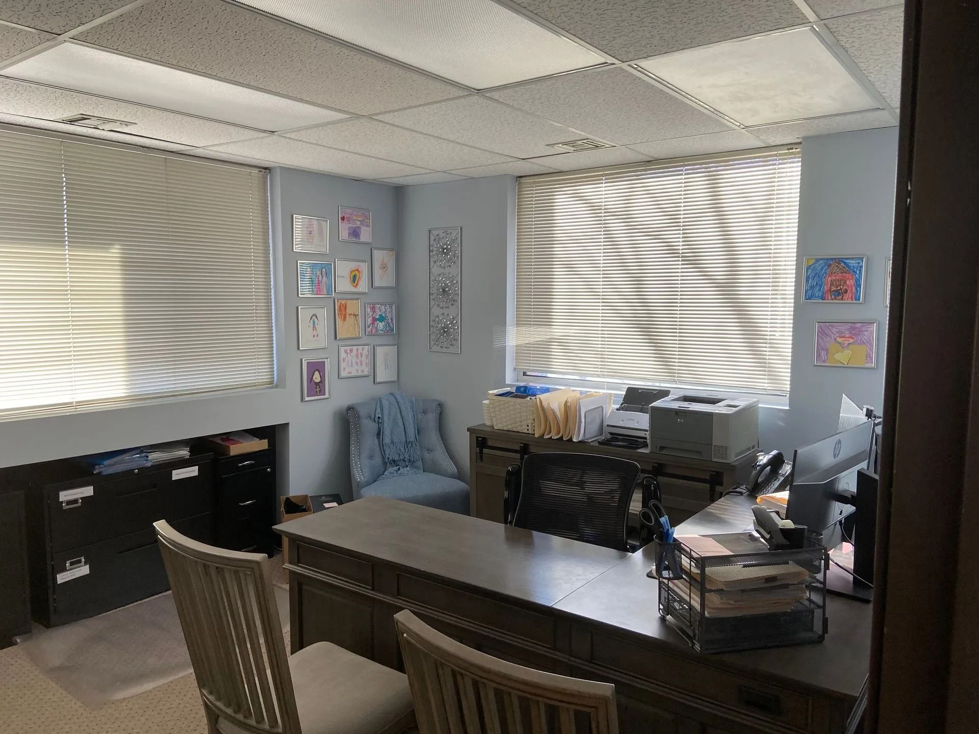 Small office with desk, chairs, file cabinets, and sunlight through window blinds.