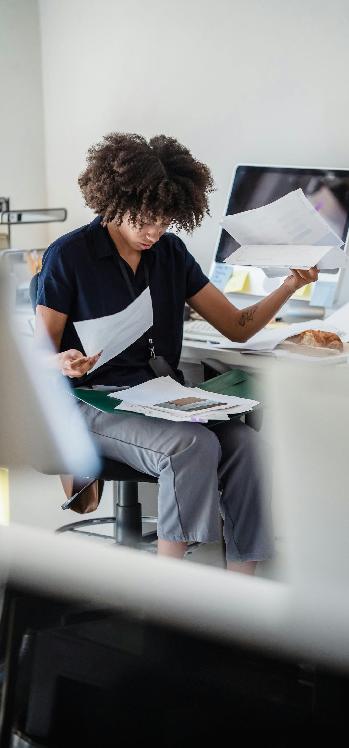 Person with curly hair, in office, reviews documents, next to a computer.