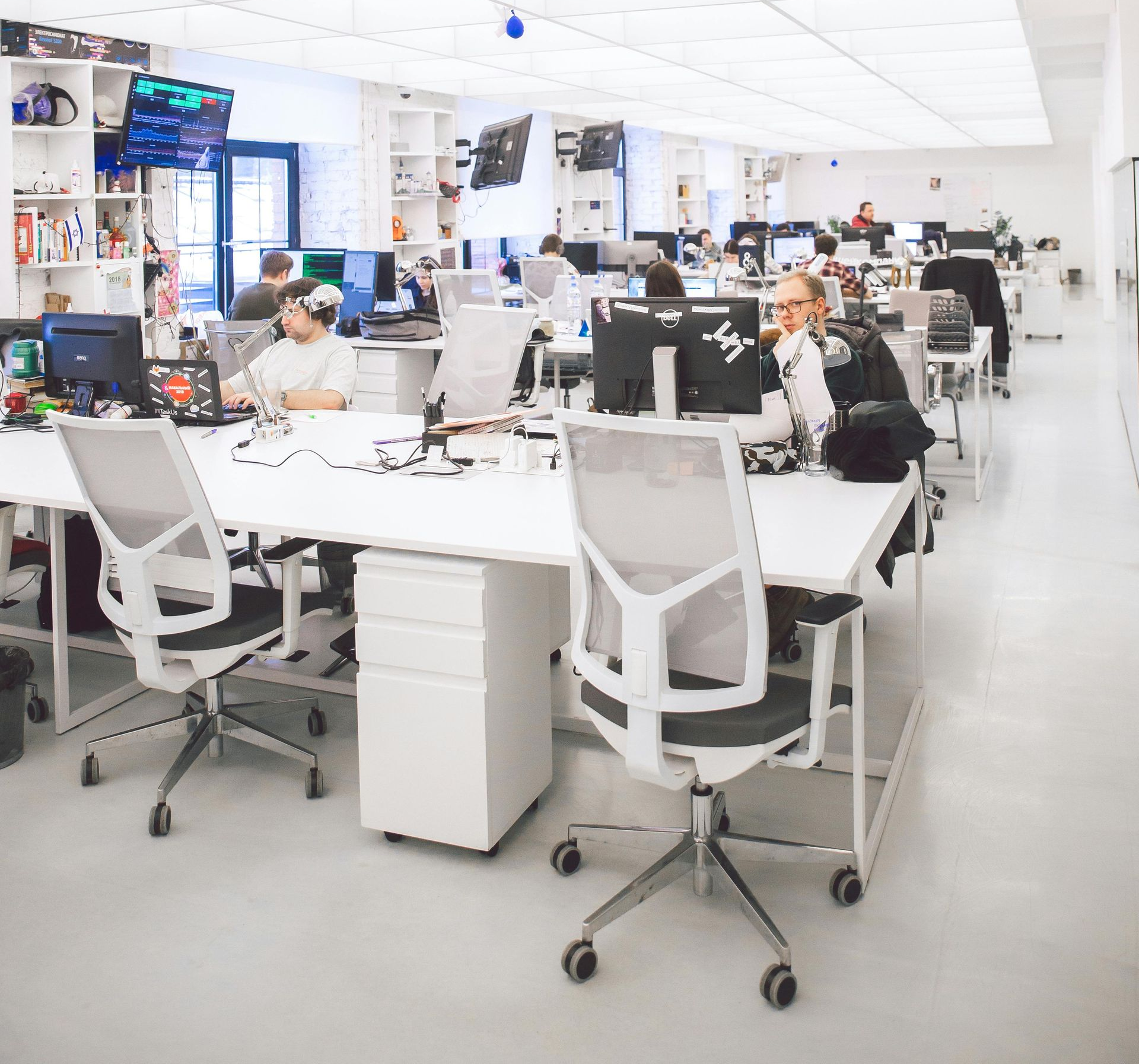 Bright, open office with multiple people working at computers. White desks and chairs, neutral color scheme.