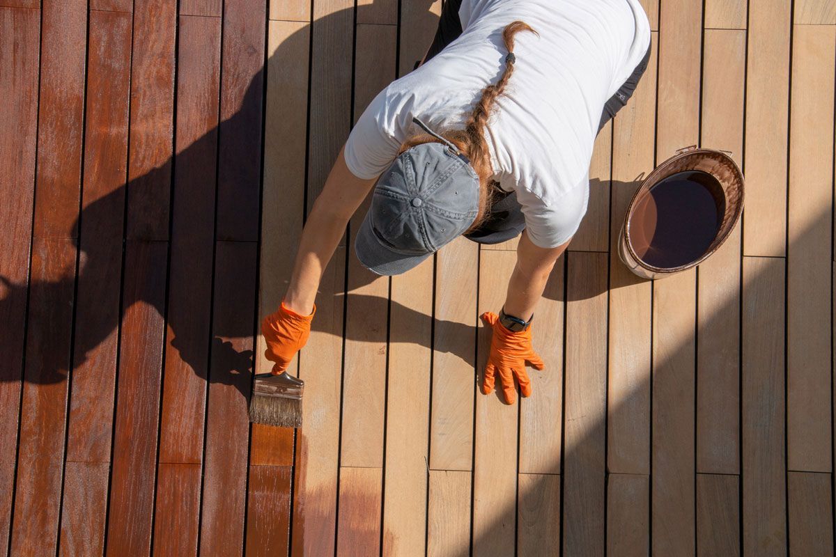 Woman staining wooden deck with brown stain, wearing hat and gloves, working outdoors.