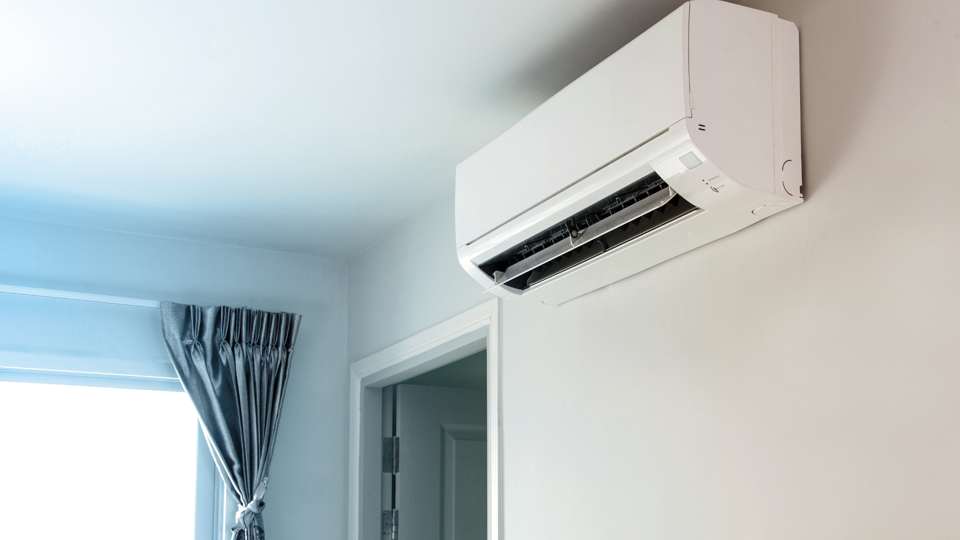 White air conditioner mounted on a white wall in a room, with a window and doorway visible.