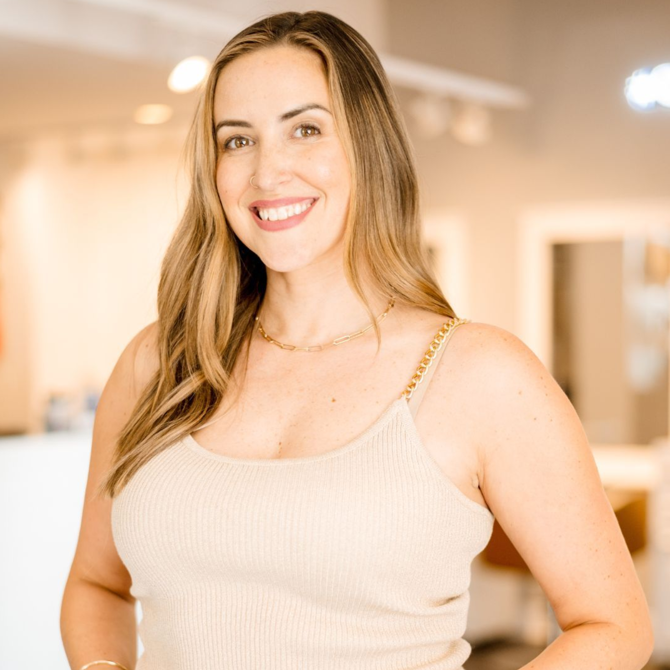 Woman smiling, tan top, gold necklace, neutral background.